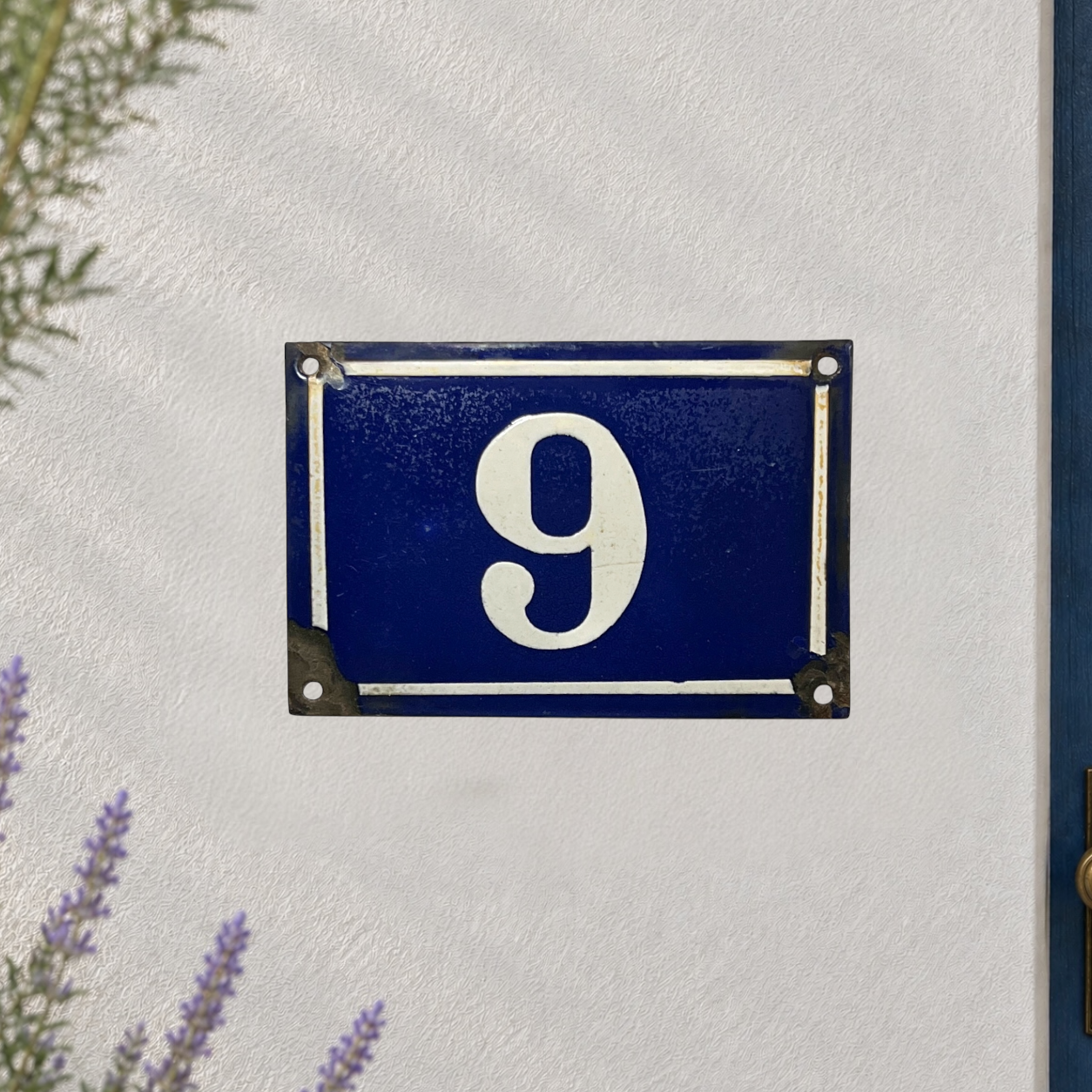 A rectangular blue house number 9 plaque with white lettering and a white border, mounted on a textured white wall with decorative greenery in the corners.