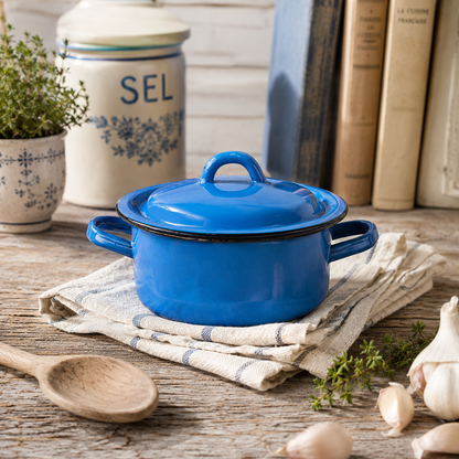 A bright blue enamel lidded pot with side handles sits on a folded cream linen cloth on a rustic wooden table. A wooden spoon, garlic, herbs, a ceramic canister labeled SEL, and books are visible in the background.