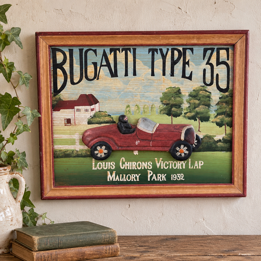 A framed vintage-style wall art print featuring a red Bugatti Type 35 race car on a painted landscape with the text 'BUGATTI TYPE 35' and 'Louis Chirons Victory Lap Mallory Park 1932.' The frame is hung on a textured cream wall above a wooden surface with books, and ivy is visible at the left edge.