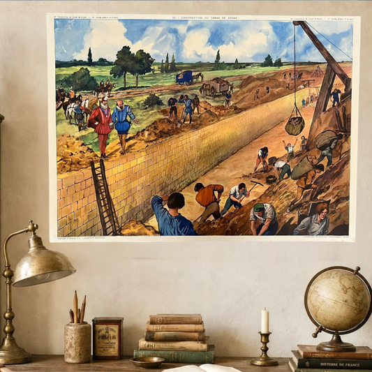 A vintage-style framed art print depicting workers building a long trench or canal, hanging on a beige wall above a desk with a lamp, books, a candle, and a globe.