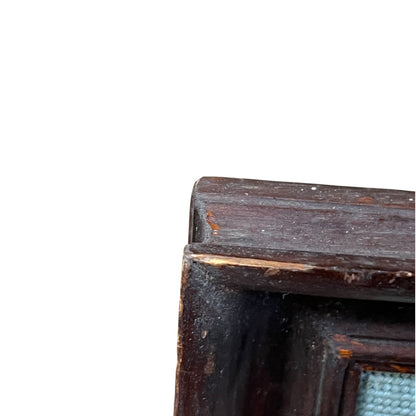 Close-up of the corner of a dark brown wooden frame or cabinet with visible wear and a small glimpse of a light blue textured insert, set against a plain white background.