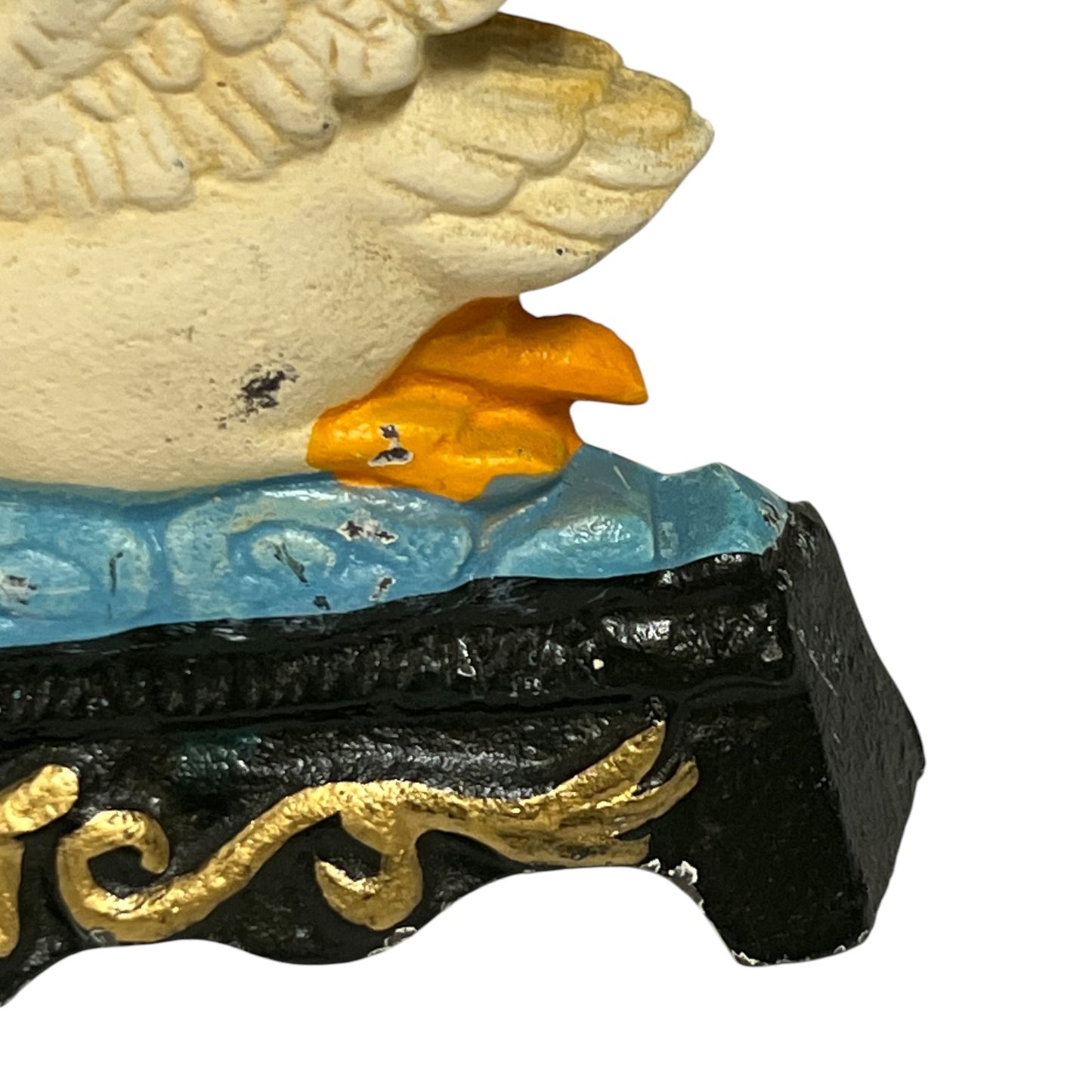 Close-up of a decorative figurine showing part of a beige bird with an orange beak above a blue wave-like surface and a black base with gold scrollwork, on a white background.