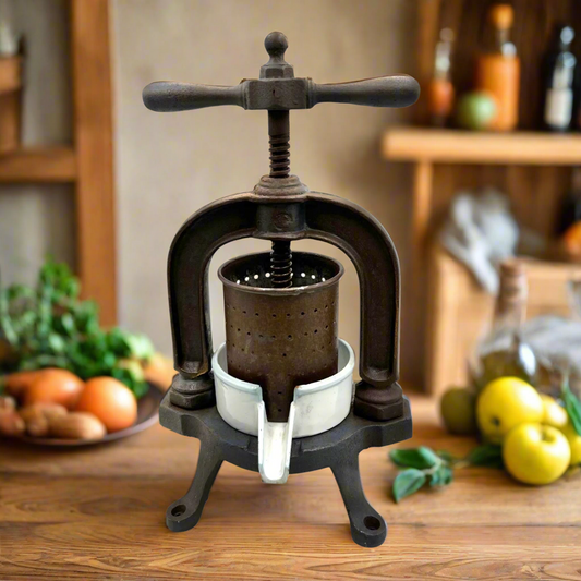Vintage French cast iron metal fruit press with a white enamel container on a kitchen table with fruit and a rustic setting in the background 