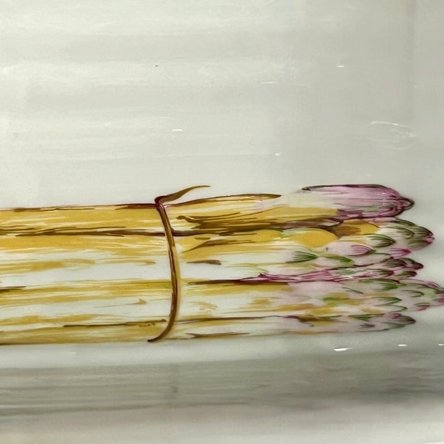 A close-up of a white ceramic plate or dish featuring a painted illustration of a bundle of asparagus tied with a brown string, with yellow-green stalks and pink-purple tips.