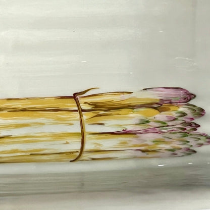 A close-up of a white ceramic plate or dish featuring a painted illustration of a bundle of asparagus tied with a brown string, with yellow-green stalks and pink-purple tips.