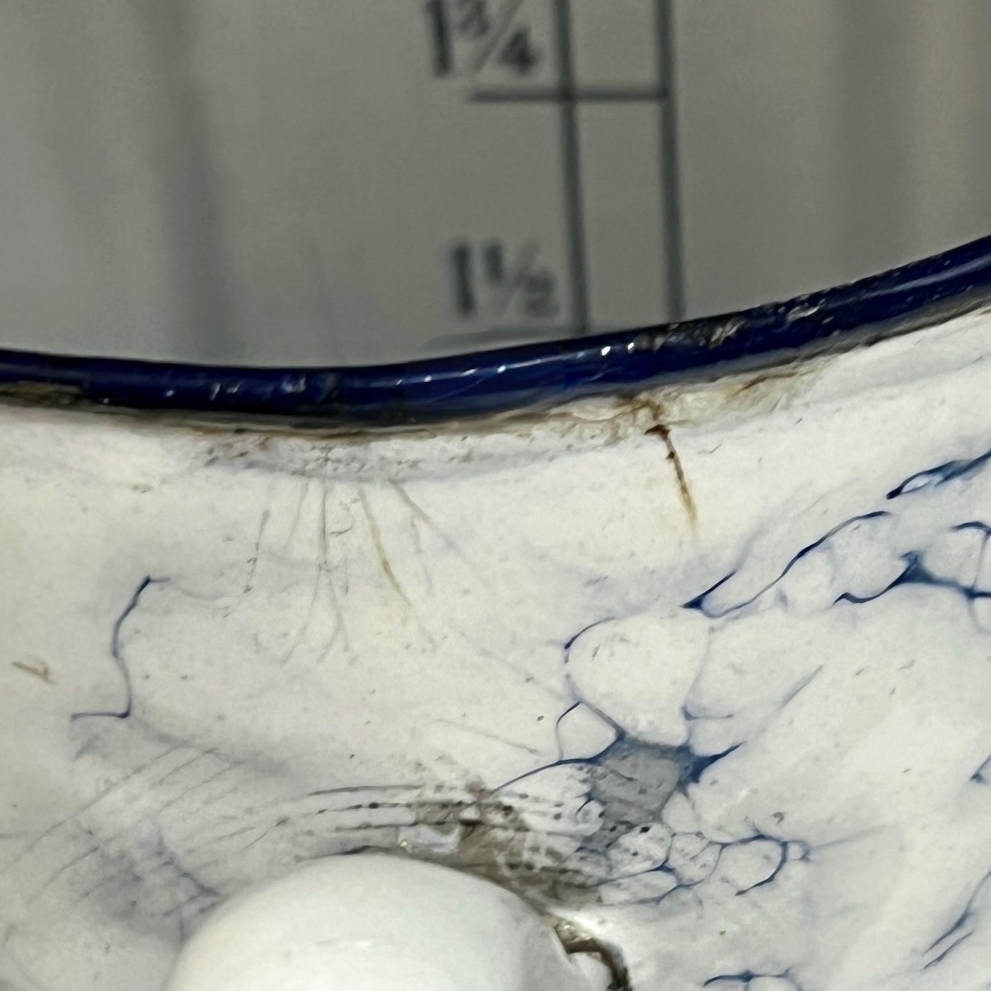 Close-up of a white pot with a dark blue rim and blue marbled pattern inside, shown against a grey background with measurement markings.