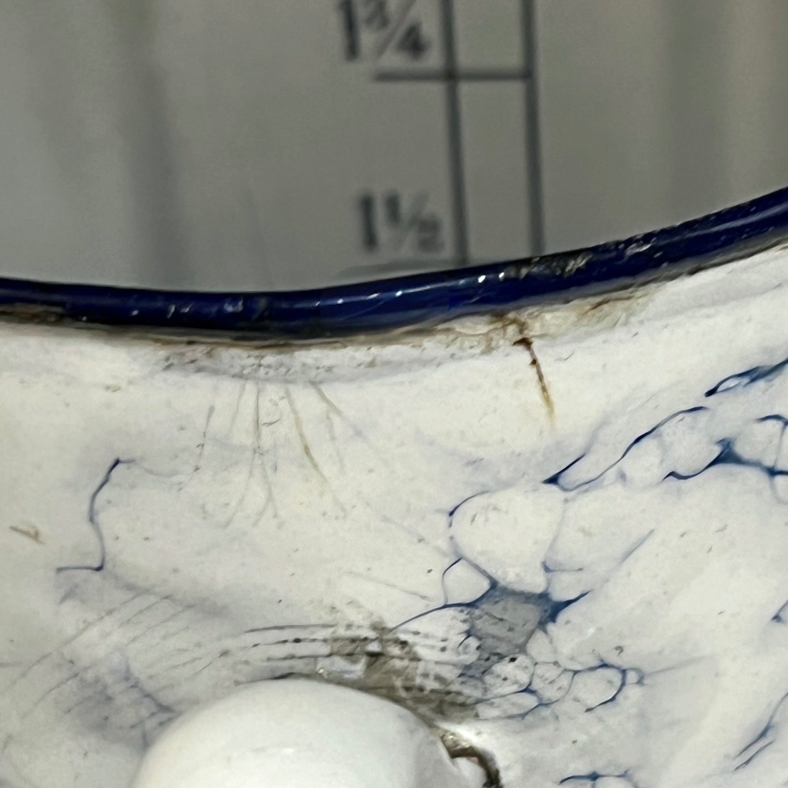 Close-up of a white pot with a dark blue rim and blue marbled pattern inside, shown against a grey background with measurement markings.