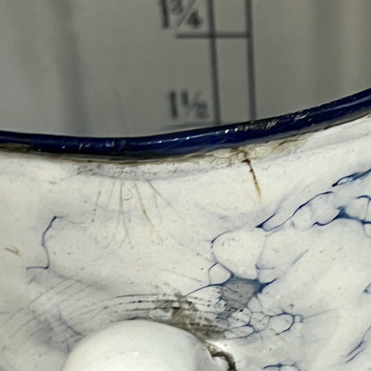 Close-up of a white pot with a dark blue rim and blue marbled pattern inside, shown against a grey background with measurement markings.