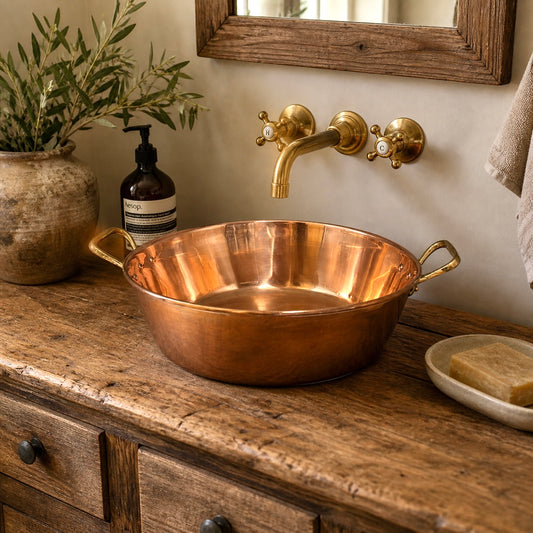 Copper up cycled basin sink with brass handles and a brass pop up waste drain  on a wooden vanity with a gold faucet, plant, and soap dish.