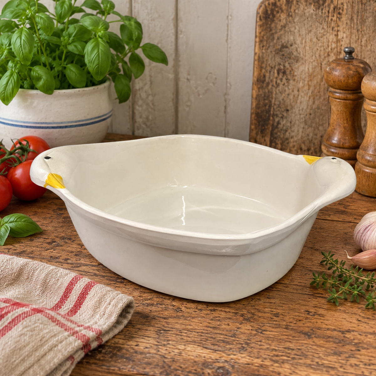 A white ceramic baking dish with small yellow handles on both sides, set on a rustic wooden table surrounded by kitchen props like tomatoes, basil, garlic, and wooden utensils.