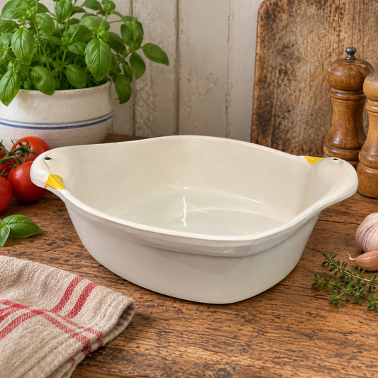 A white ceramic baking dish with small yellow handles on both sides, set on a rustic wooden table surrounded by kitchen props like tomatoes, basil, garlic, and wooden utensils.