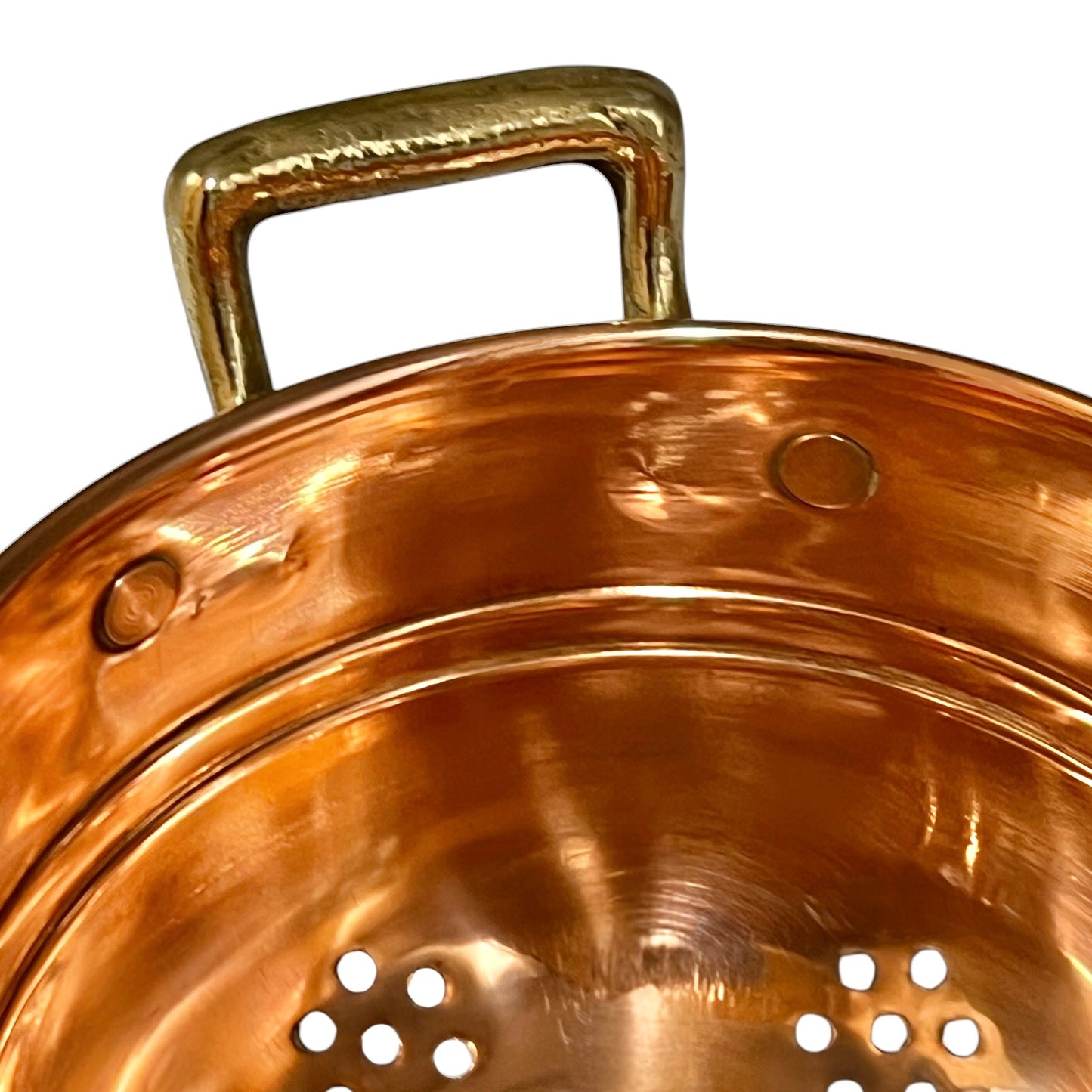 Close-up of a copper pot or colander with a hammered brass handle, showing the reflective interior and perforated holes on the bottom against a white background.