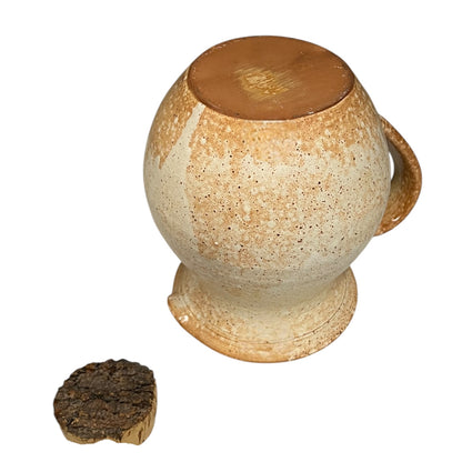 Ceramic jug with a textured surface and handle, next to a small piece of dark material on a white background.