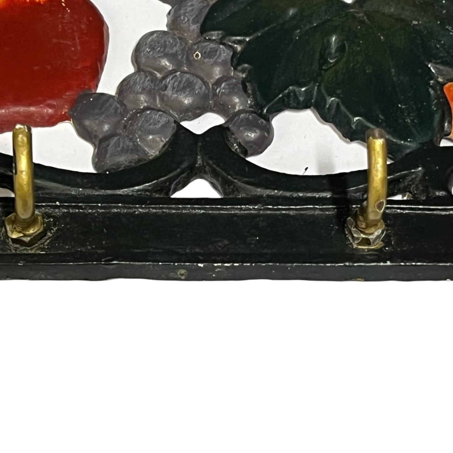 Close-up of a decorative wall hooks showing dark green leaves, gray grape clusters, a red fruit shape, and a black frame with brass hooks against a white background.