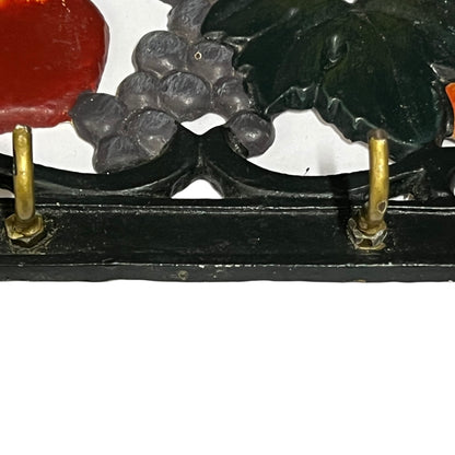 Close-up of a decorative wall hooks showing dark green leaves, gray grape clusters, a red fruit shape, and a black frame with brass hooks against a white background.
