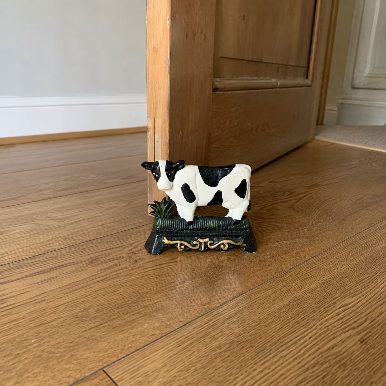 A small cast iron doorstop shaped like a cow, perfect for adding charm to a country cottage or farmhouse, displayed on a wooden floor with a door ajar in the background.