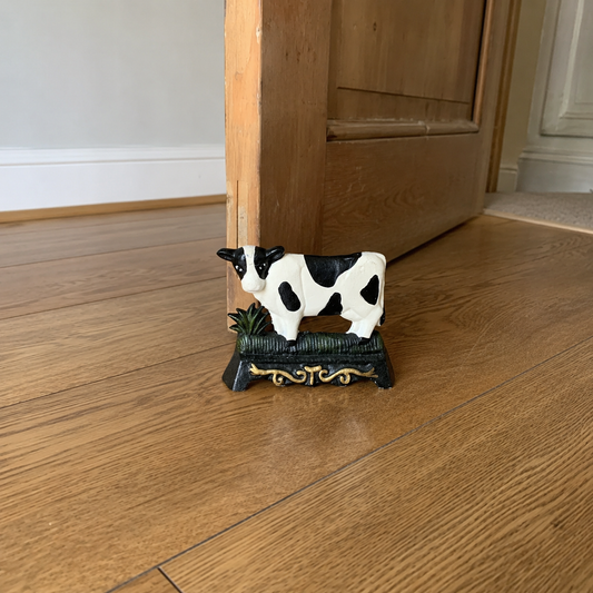 A small cast iron doorstop shaped like a cow, perfect for adding charm to a country cottage or farmhouse, displayed on a wooden floor with a door ajar in the background.