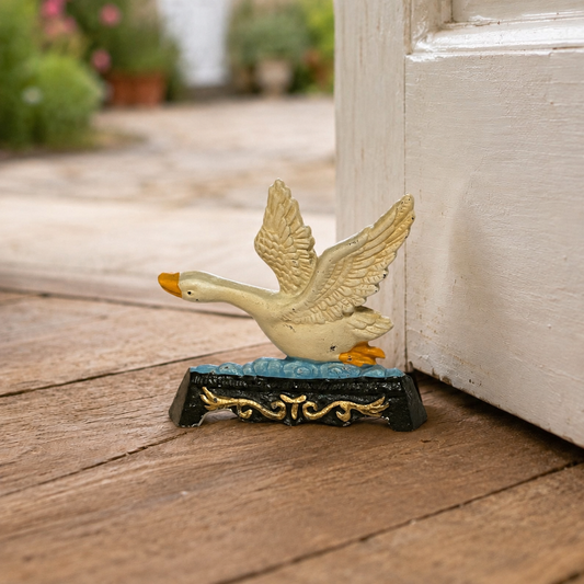 A decorative goose figurine with outstretched wings standing on a black and gold base, placed on a wooden surface beside a white door in an outdoor setting.