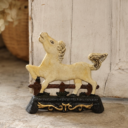 A decorative cast iron horse doorstop with a cream-colored body and wings, standing on a dark base with gold scrollwork, set against a rustic white wooden background.