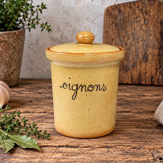 A speckled yellow ceramic canister with a lid and the word 'oignons' written in black script, set on a rustic wooden table with kitchen props including a garlic bulb, herbs, and a cutting board.