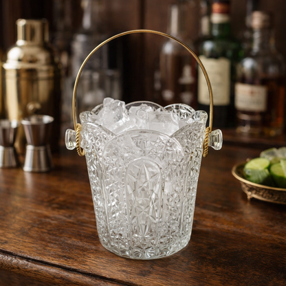 Decorative glass ice bucket with gold handle on a wooden bar counter with bottles and bar tools in the background.