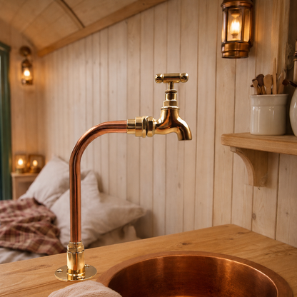 A copper faucet with a cross handle and matching copper sink basin set into a rustic wooden countertop, with a softly blurred bedroom background and warm wall sconces.