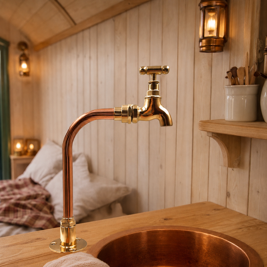 A copper faucet with a cross handle and matching copper sink basin set into a rustic wooden countertop, with a softly blurred bedroom background and warm wall sconces.