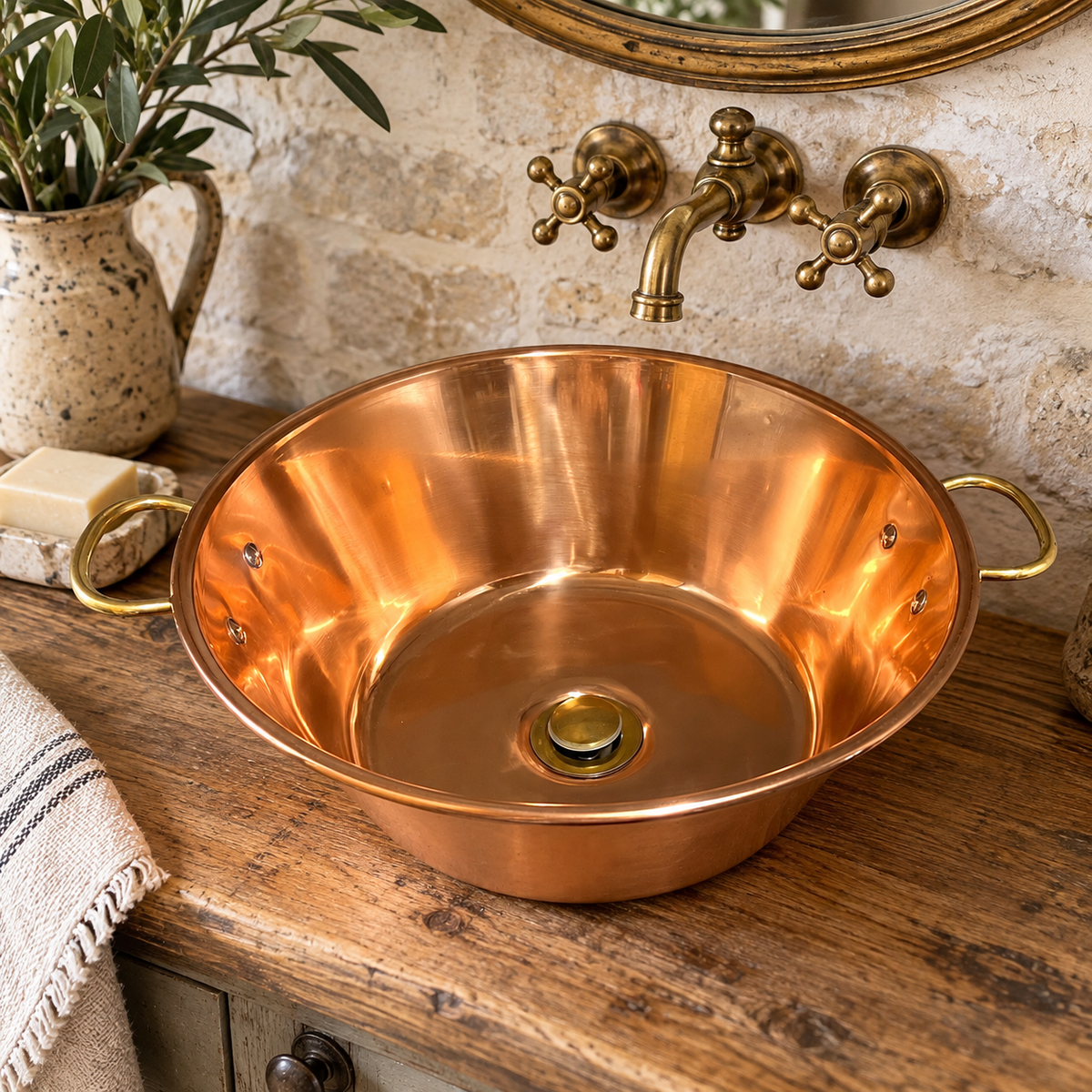 A copper vessel sink with side handles sits on a rustic wooden bathroom counter beneath an antique brass faucet, with a stone wall, mirror, soap dish, and small plant in the background.