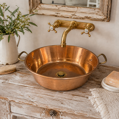 A round copper basin sink with a matching brass faucet and cross handles, set on a distressed wooden vanity with a vase, soap dish, and towel nearby.