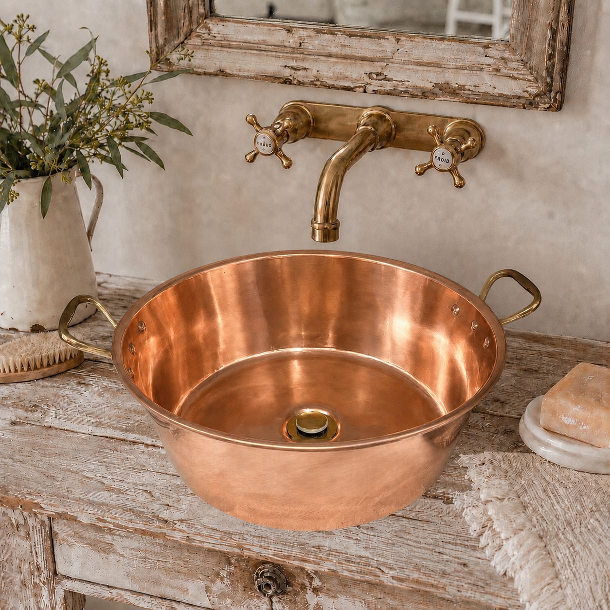 A round copper bathroom sink with a matching brass faucet and handles, mounted on a distressed wooden vanity with decorative props including a vase of greenery and soap.