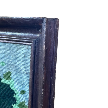 Close-up of the corner of a framed needlepoint canvas, showing a dark stained wooden frame and light blue canvas with green and dark green stitched details, set against a plain white background.