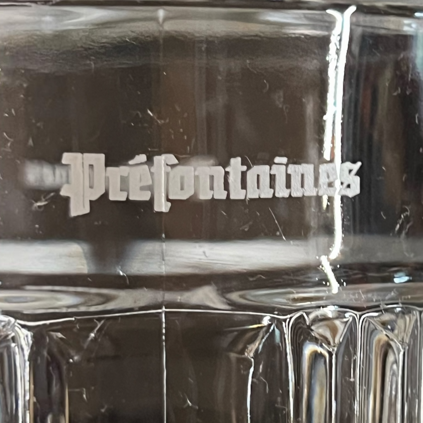 Close-up of a clear glass mug or tumbler with the word 'Préfontaines' printed in white gothic-style lettering on the side.