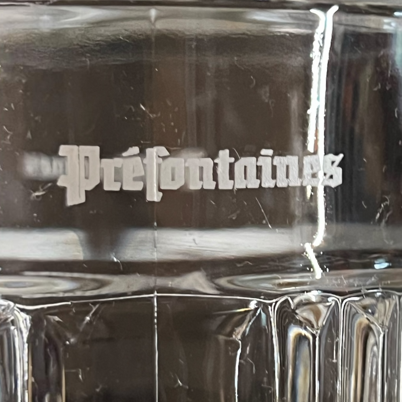 Close-up of a clear glass mug or tumbler with the word 'Préfontaines' printed in white gothic-style lettering on the side.