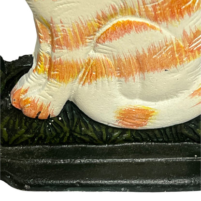 Close-up of a hand-painted cat figurine with orange and yellow stripes on an off-white body, sitting on a dark textured base.