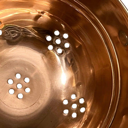 Close-up of the inside of a copper pot or steamer basket, showing a shiny metallic copper surface with multiple circular drainage holes and a reflective finish.
