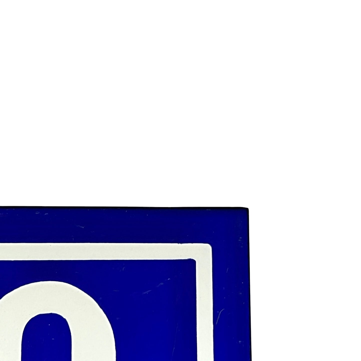 A close-up of a blue and white sign corner showing a white letter inside a blue square, with most of the frame filled by a plain white background.