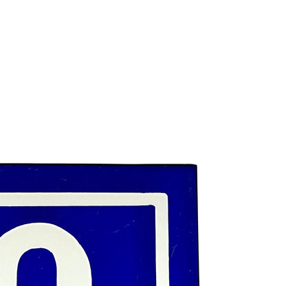 A close-up of a blue and white sign corner showing a white letter inside a blue square, with most of the frame filled by a plain white background.
