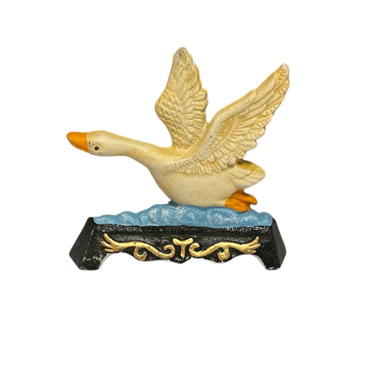 A decorative cast iron duck figurine in flight, featuring a cream-colored body and wings with orange beak and feet, standing on a blue and black base with gold scrollwork, isolated on a white background.