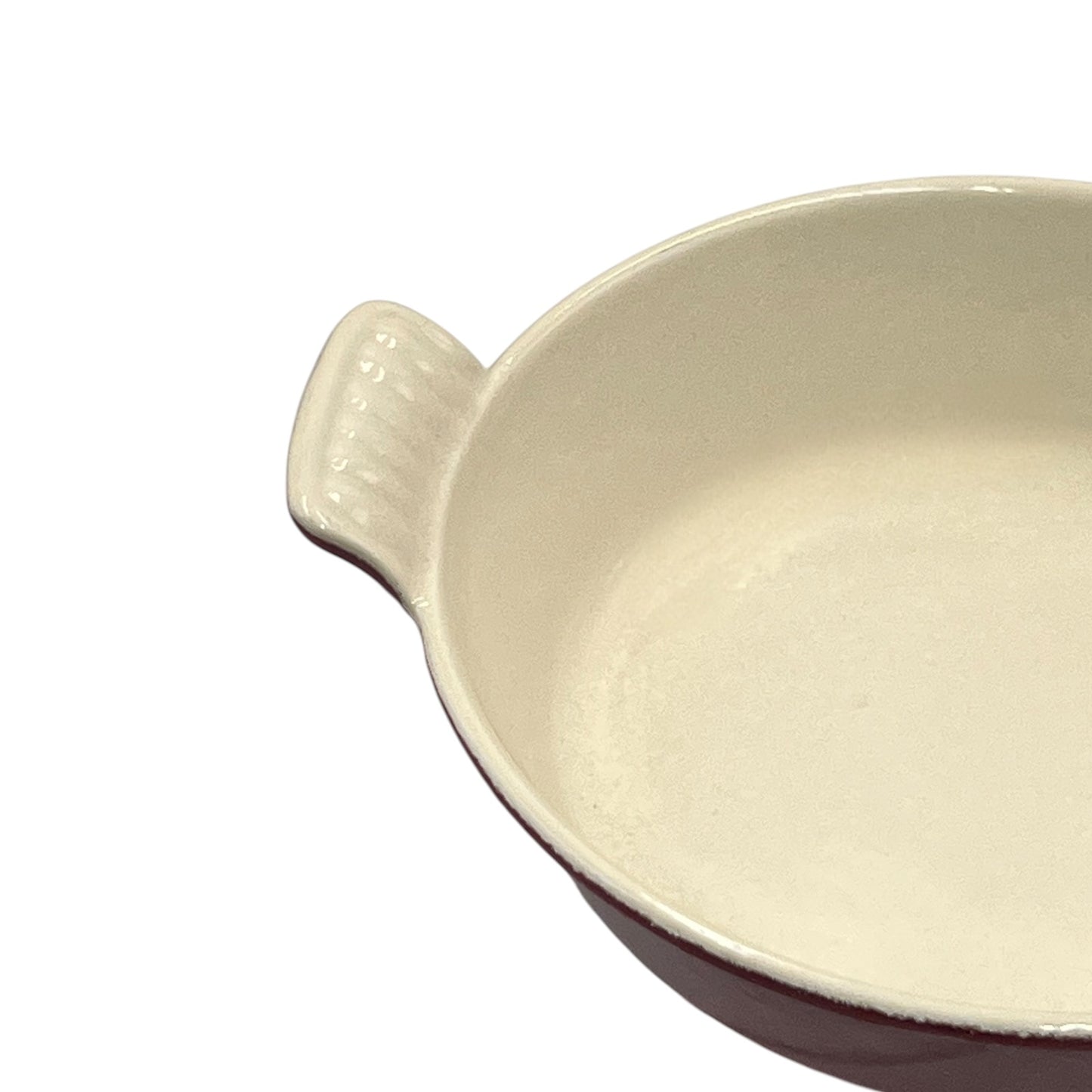 A close-up of a ceramic baking dish with a cream-colored glazed interior and a dark reddish-brown exterior, featuring a small ribbed handle on the left side, set against a plain white background.