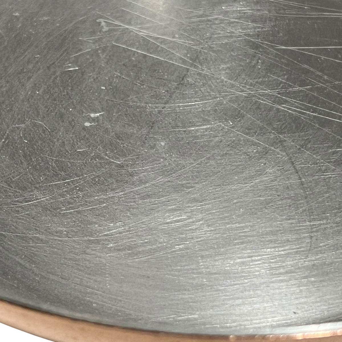 Close-up of a scratched dark gray metallic surface, likely a pan or cookware interior, with visible scratches and a small copper-toned rim at the bottom edge.