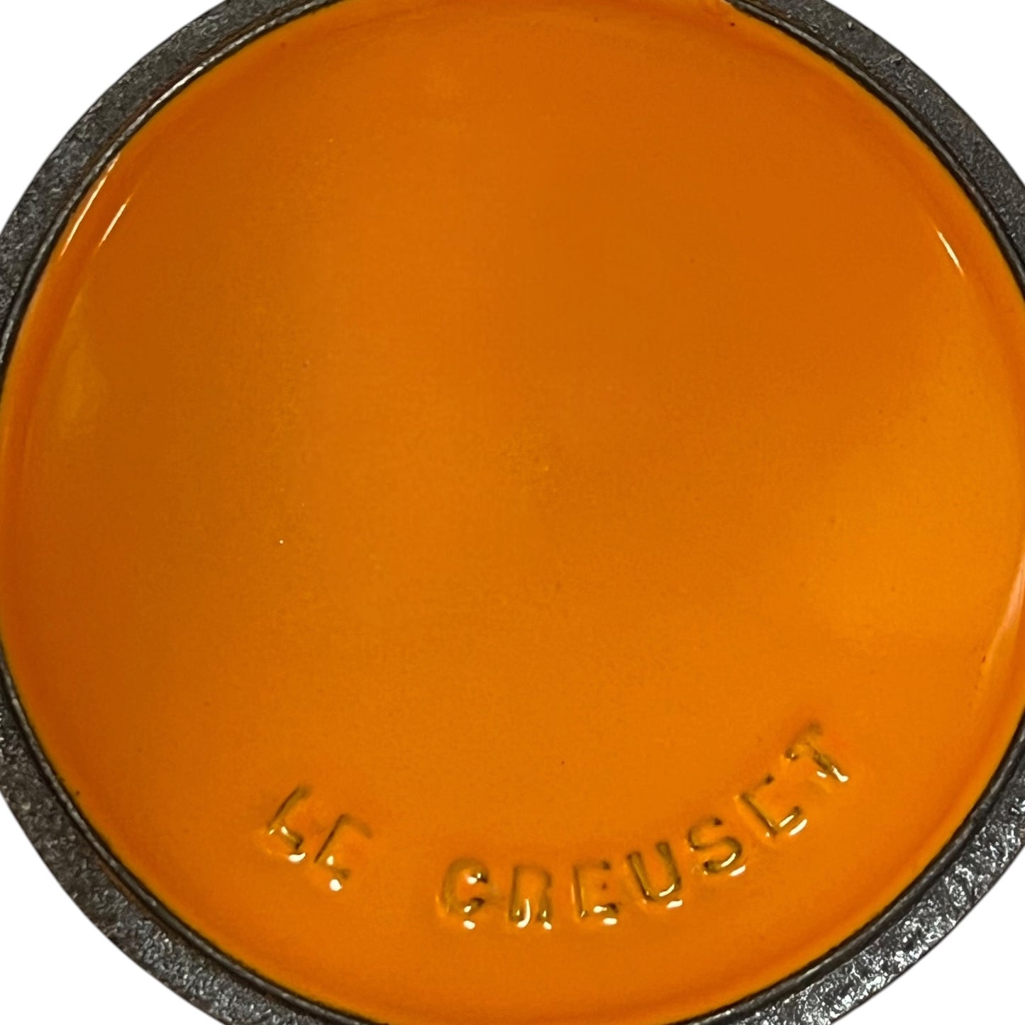 A close-up of a round orange color swatch with the brand name 'LE CREUSET' embossed at the bottom, surrounded by a dark textured rim.