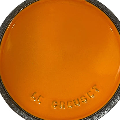 A close-up of a round orange color swatch with the brand name 'LE CREUSET' embossed at the bottom, surrounded by a dark textured rim.