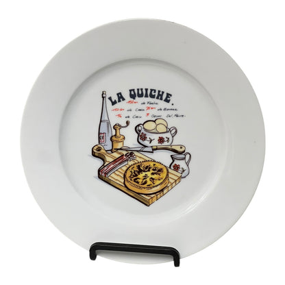 White plate with 'La Quiche' design featuring a pie, bottle, and other items on a stand.