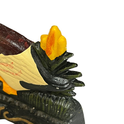 Close-up of a decorative door stop or sculpture showing a dark green leafy form, a cream and red side panel, and a bright yellow-orange flame-like top detail on a plain white background.