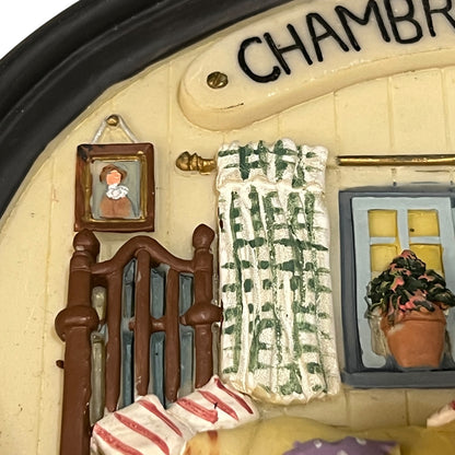 Close-up detail of a miniature decorative figurine or room scene sign showing part of a cream-coloured wall with black lettering reading "CHAM.