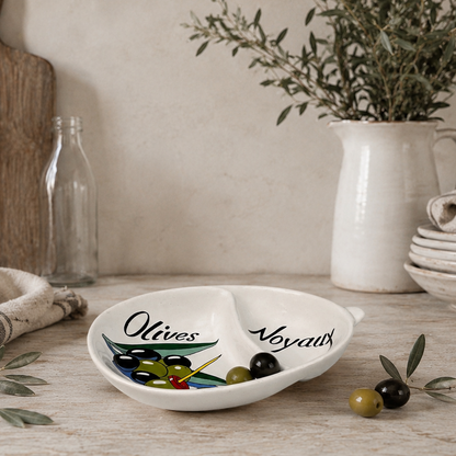 Decorative dish with olive design on a wooden surface with plants and kitchen items.