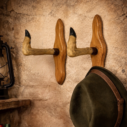 Two rustic wooden wall hooks made from taxidermy deer hooves, mounted on a textured stone wall. A dark green hat with a brown leather strap hangs from the right hook, and part of a black lantern is visible on the left.