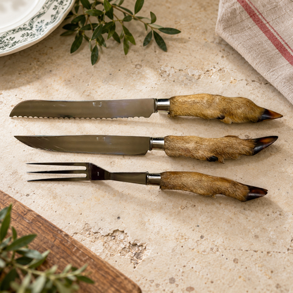 A three-piece knife set with animal-hide handles, including a serrated steak knife, a dinner knife, and a carving fork, arranged on a textured stone surface with rustic props.
