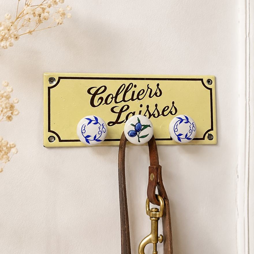 A rectangular cream-colored metal wall hook sign with black script reading 'Colliers Laisses' and black border lines, mounted on a light wall. Three round ceramic hooks with blue floral designs hang below, along with a brown leather strap and brass clasp.