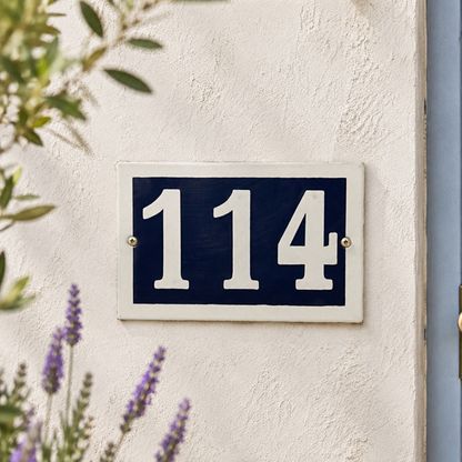 House number '114' on a blue and white sign against a light wall with plants in the background.